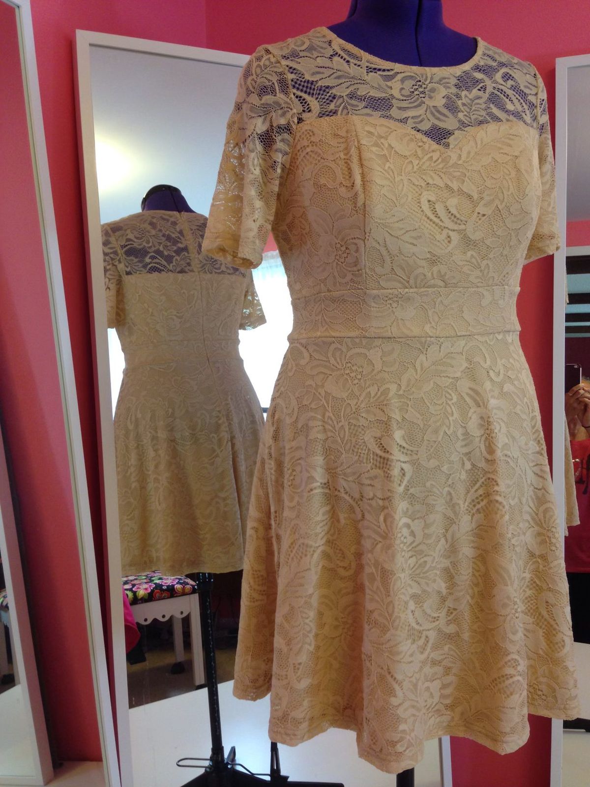 A gold lace knee-length dress with a sheer lace neckline on a dress form, mirrors behind